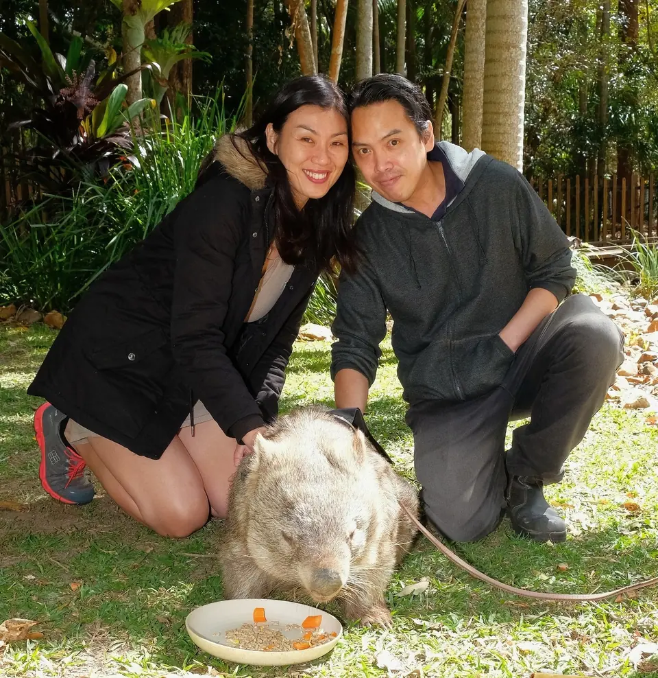 Paddy Picho and his wife crouched on the grass beside a wombat eating from a bowl, at Australia Zoo in Queensland. Paddy is on the right.